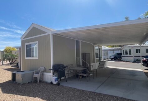 A small beige mobile home with a covered carport, outdoor table and chairs, grill, and nearby parked cars under a clear blue sky.