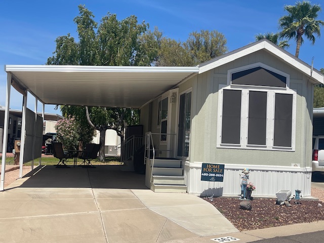 A manufactured home with a covered carport, front steps, and a "Home for Sale" sign, situated in a neighborhood with palm trees and other similar homes nearby.