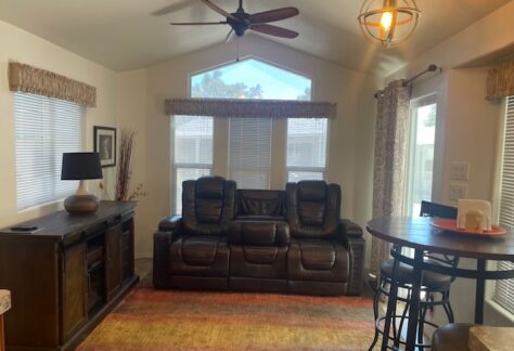 A living room with a brown leather reclining sofa, a wooden cabinet with a lamp, a round dining table with stools, and large windows letting in natural light.