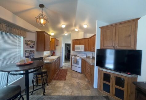 A kitchen with wood cabinets, white appliances, tiled floor, a round dining table with two chairs, and a flat-screen TV on a cabinet. Ceiling lights and a window provide bright lighting.