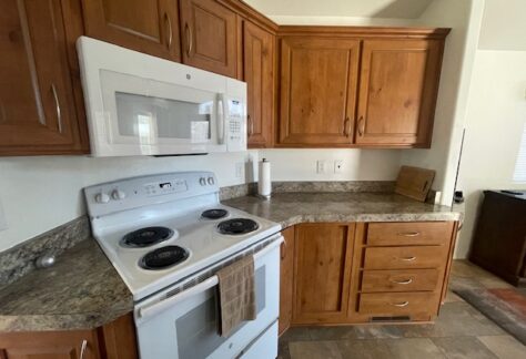 Kitchen with wooden cabinets, an electric stove with a towel, a microwave, paper towel holder, and a cutting board on a marble-patterned countertop.