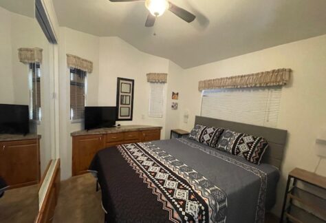 A bedroom with a neatly made bed, patterned bedding, wooden cabinets, a flat-screen TV, and windows with blinds and valances. A ceiling fan is on the white ceiling.