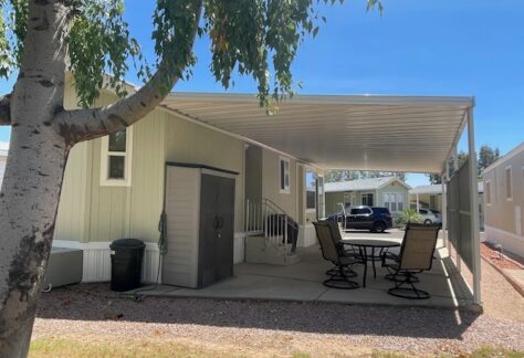 A covered patio with four chairs and a table beside a light green mobile home, with a tree in the foreground and other homes visible in the background.