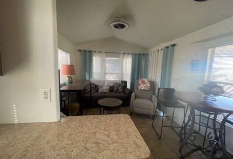 Small living room with a couch, armchair, side table, bar-style dining table, and stools. Large windows with curtains let in natural light. Countertop in the foreground.