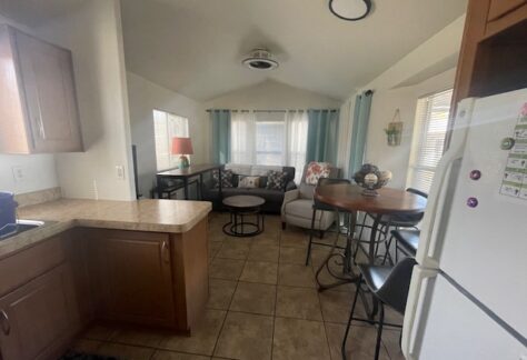 Small living area with tiled floor, a kitchen on the left, a round dining table, and a seating area with a sofa, chairs, and a lamp near windows with light blue curtains.