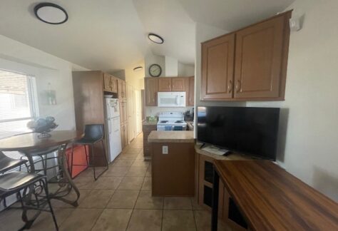A kitchen with brown cabinets, white appliances, a wall clock, a small island, a round dining table with chairs, and a TV on a wooden desk.