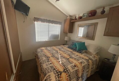 Small bedroom with patterned bedspread, pillows, two lamps, wall-mounted TV, a window with blinds, and decorative items on a shelf above the bed.