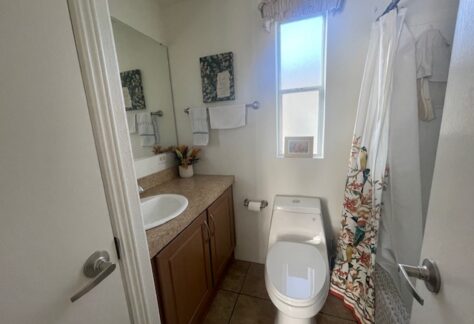 A small bathroom with a sink, countertop, toilet, mirror, shower with a curtain, towel racks, and a window letting in natural light.
