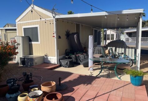 A small beige mobile home with a covered patio, outdoor furniture, potted plants, and various items arranged on the ground, under a clear blue sky.