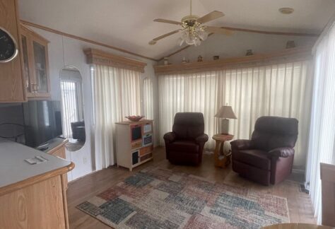 Spacious room with two leather recliners, a wooden side table with a lamp, light-colored curtains, ceiling fan, patterned area rug, and built-in wooden cabinets.