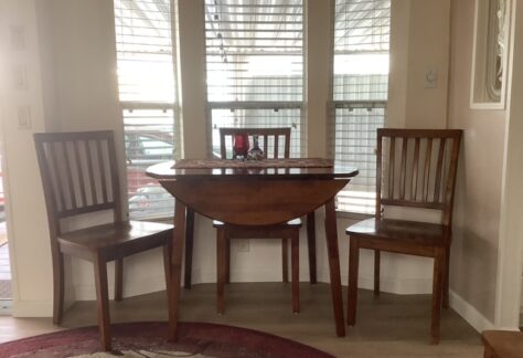 Small wooden dining table with two matching chairs is set by a bay window. A decorative centerpiece and placemat are on the table. Light streams in through the blinds.