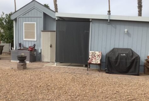 A small gray shed with a covered grill, a floral chair, storage cabinet, and paver patio, set on a gravel yard.