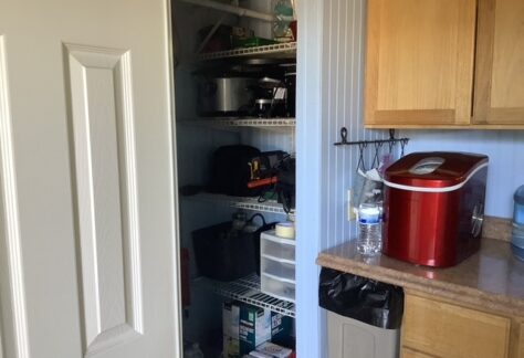 A pantry with wire shelves holding kitchen items and appliances, next to a counter with a red ice maker, water bottle, and trash can below.