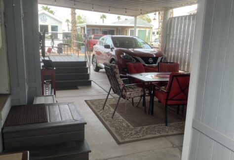 Covered patio area with a dining table and chairs on a rug, adjacent to a maroon SUV parked in a driveway; houses are visible in the background.