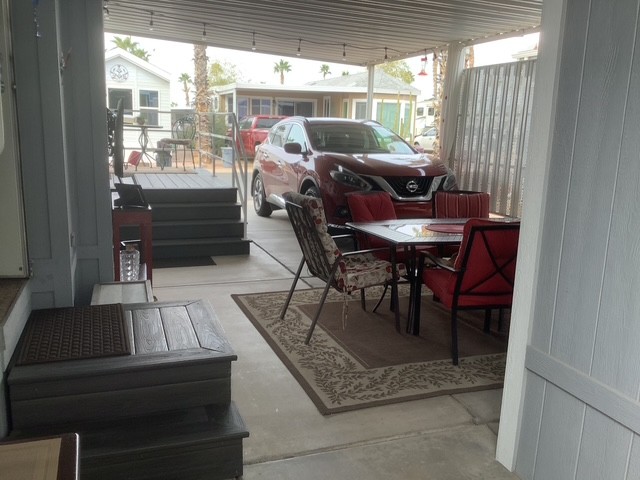 Covered patio with a table and four chairs on a rug, next to a parked red crossover SUV, and a small set of steps leading to a raised area.