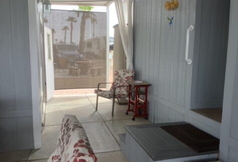 Covered patio area with gray walls, two patterned chairs, a small red table, and white curtains. Outdoor sunlight and palm trees visible through a mesh screen.