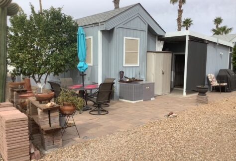 Small gray house with a patio area, outdoor dining set, barbecue grill, potted plants, and a shed, surrounded by gravel and palm trees.