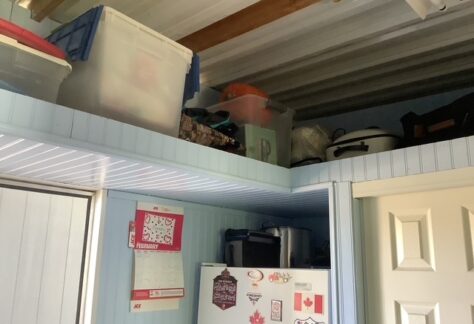 Plastic storage bins, bags, and other items are stored on a high shelf above a white refrigerator and a door in a room with light blue paneling.