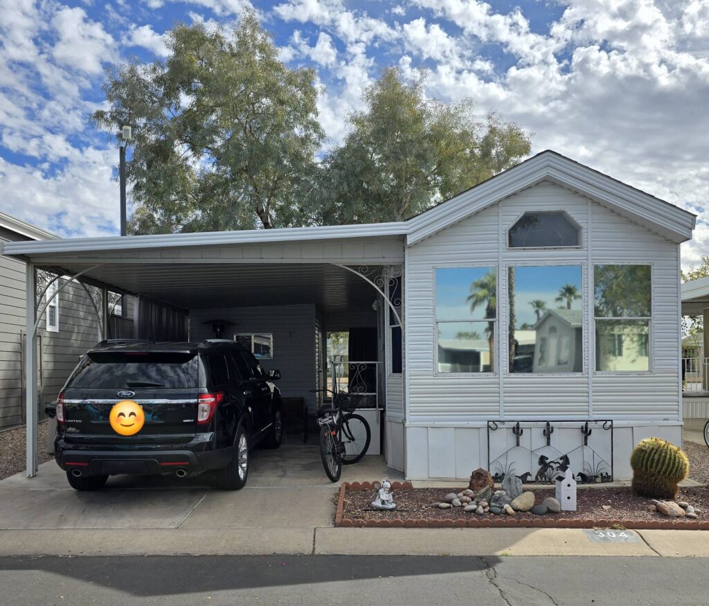 A black SUV parked under a carport beside a white mobile home. A bicycle is next to the car. Desert landscaping with rocks and small plants is in the foreground.