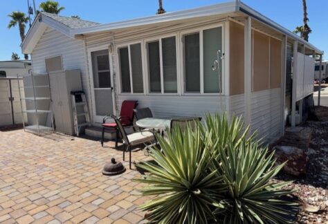 Small white mobile home with an enclosed porch, outdoor seating area, and desert vegetation, including a spiky green plant, set on a brick patio under a clear blue sky.