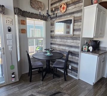 A small dining area with a round black table and four chairs. Wooden accent wall with a mirror, cabinets on the right, and a chandelier above. Neutral-toned decor and rug on the floor.