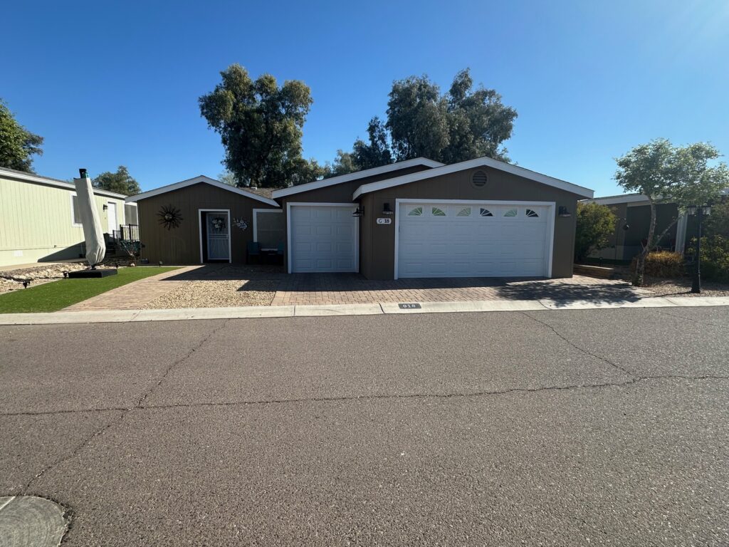 Single-story house with gray siding, two white garage doors, and a paved driveway. The yard is landscaped with gravel and a small patch of artificial grass, under a clear blue sky.