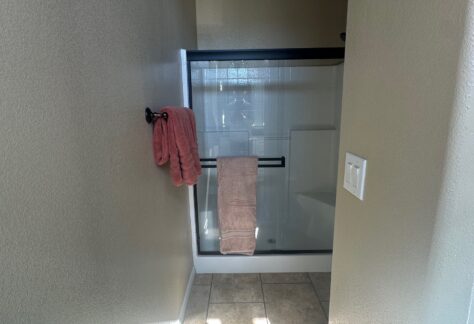 Narrow bathroom with a tiled floor, a shower with sliding glass doors, and pink towels hanging on a black bar. Natural light enters from a window above the shower.