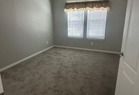 Empty bedroom with gray carpet, light gray walls, ceiling fan, two windows with blinds and a valance, viewed from the doorway.