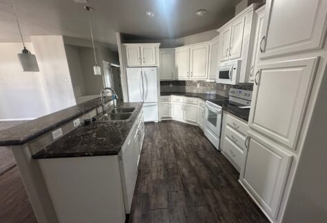 Modern kitchen with white cabinets, dark countertops, stainless steel appliances, double sink, and wood-look flooring, viewed from the corner near the breakfast bar.