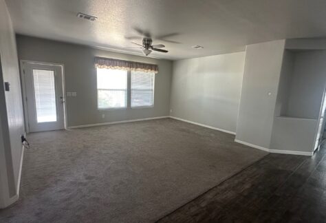 Empty living room with gray carpet, ceiling fan, two large windows with a valance, a door to the left, and dark wood flooring leading to another area on the right.