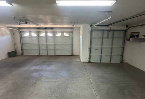 A clean, empty garage with two closed garage doors, concrete floor, ceiling lights, and a small shelf with a framed photo and object on the right wall.