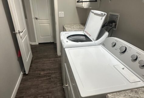 A laundry room with a washing machine and dryer, both white, side by side under a hanging rod and shelves. The washing machine lid is open. The floor is dark wood.
