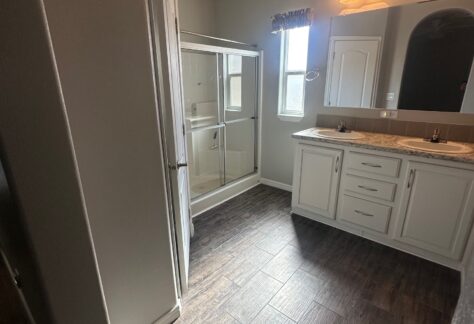 Bathroom with tiled floor, white cabinets, a double sink vanity with mirrors, a glass shower enclosure, and a window allowing natural light.