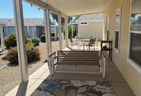 A covered patio with a floral rug, wooden porch swing, table with chairs, and desert landscaping in the background.