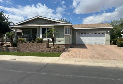 Single-story house with a front porch, two-car garage, brick driveway, small palm trees, and landscaped yard under a partly cloudy sky.