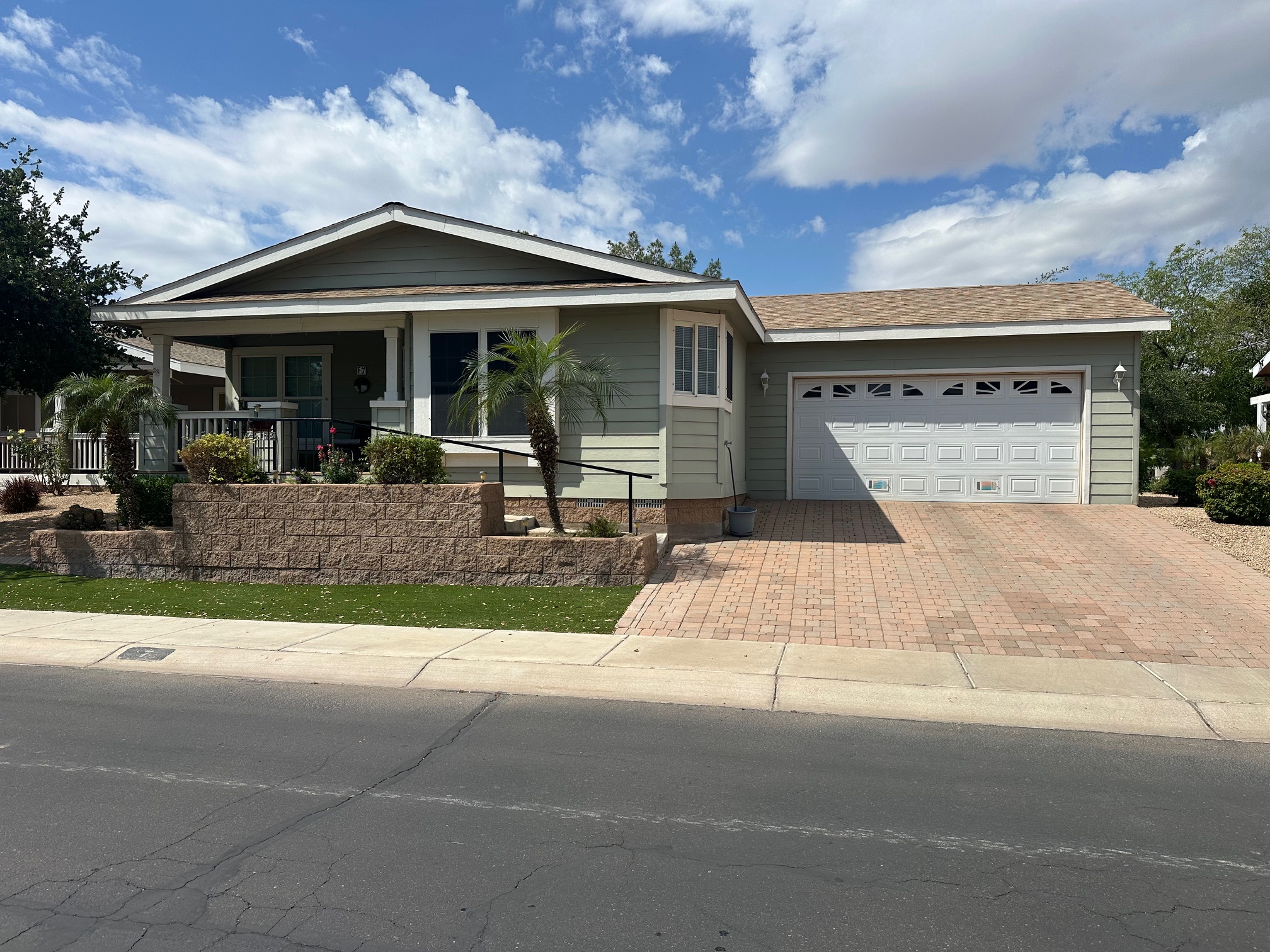 Single-story house with a front porch, attached two-car garage, brick driveway, and landscaped yard with small palm trees and shrubs, under a partly cloudy sky.