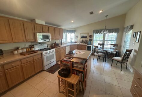 Spacious kitchen with light wood cabinets, a gas stove, center island with stools, and a dining area by large windows with white tile flooring.
