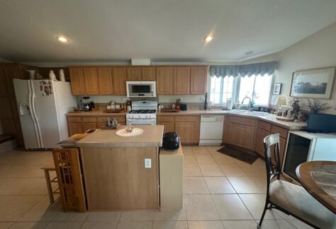 A spacious kitchen with wooden cabinets, an island, white appliances, and a small round dining table with two chairs near a window.