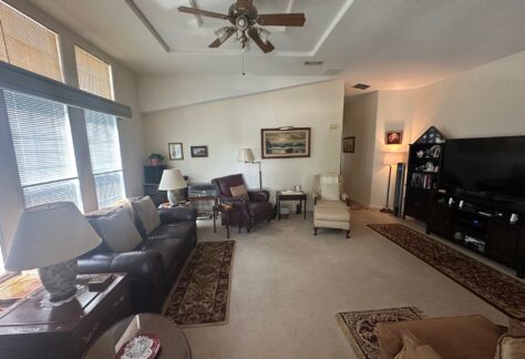 A living room with sofas, armchairs, rugs, a ceiling fan, multiple lamps, a TV, and wall art, featuring light carpeted flooring and large windows with blinds.
