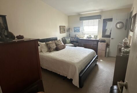 A small, neatly arranged bedroom with a double bed, dark wood furniture, decorative items, and a window letting in natural light.