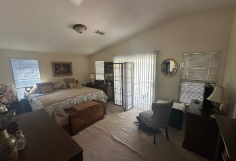 A bedroom with a large bed, two dressers, a chair at a desk, a folding room divider, and sliding glass doors with vertical blinds letting in natural light.