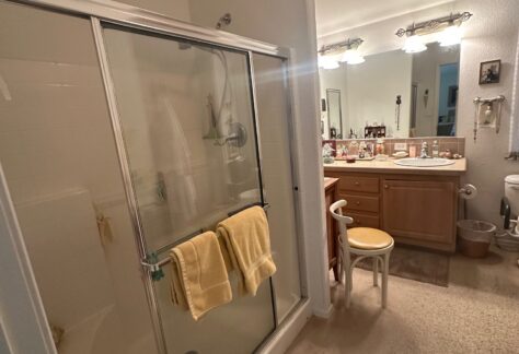 A bathroom with a glass shower, two yellow towels hanging on the door, a sink with a mirror, and various toiletries on the counter. Carpet covers the floor.