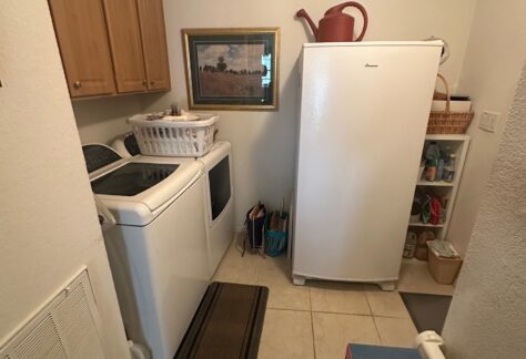 Laundry room with a washer, dryer, and upright freezer. Cabinets, baskets, cleaning supplies, and a framed picture are visible. The floor is tiled and the room is well lit.