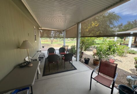 Covered patio area with several chairs around a small mat, a side table with a lamp, and potted plants, overlooking a landscaped yard and trees in the background.