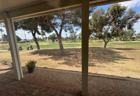View from a covered patio overlooking a grassy golf course with golf carts, trees, and a partly cloudy sky in the background.