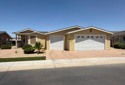 Single-story yellow house with two attached garages, brick driveway, desert landscaping, and a clear blue sky in the background.