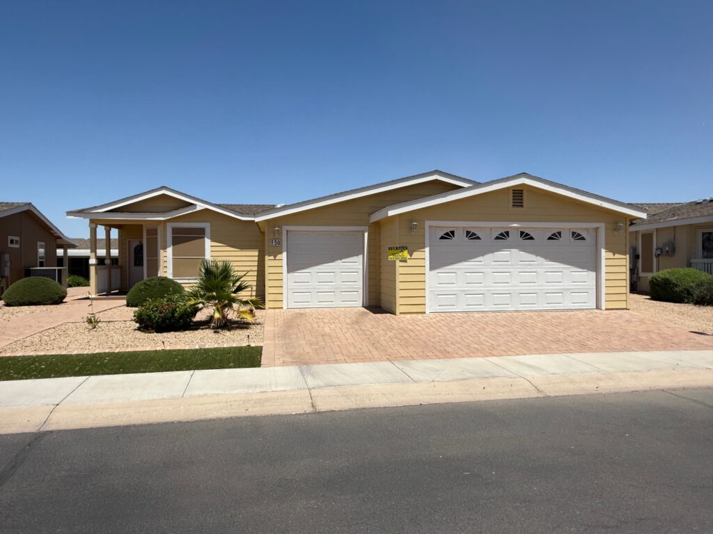 Single-story yellow house with a front porch, manicured bushes, and a three-car garage, situated on a paved driveway under a clear blue sky.