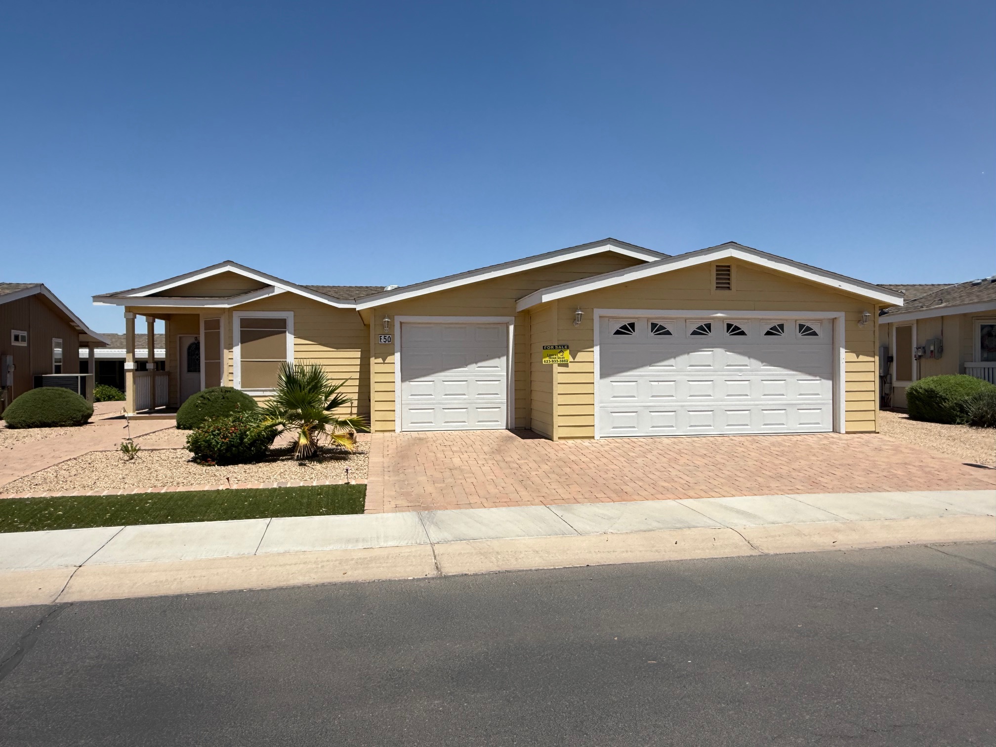 Single-story yellow house with a front porch, manicured bushes, and a three-car garage, situated on a paved driveway under a clear blue sky.